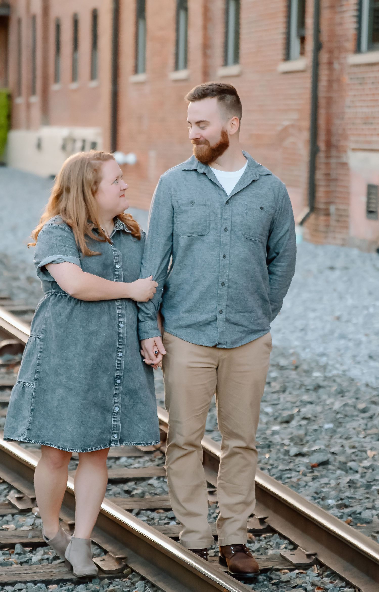 A couple in coordinated denim and khaki outfits stands together on railroad tracks in multiple poses during a photoshoot.