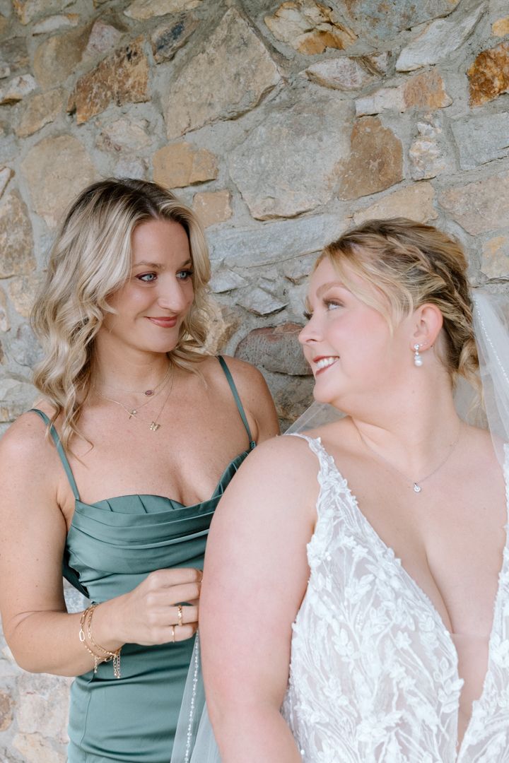 Two people in formal attire share candid moments in front of a stone wall, one wearing sage green and one in white lace.