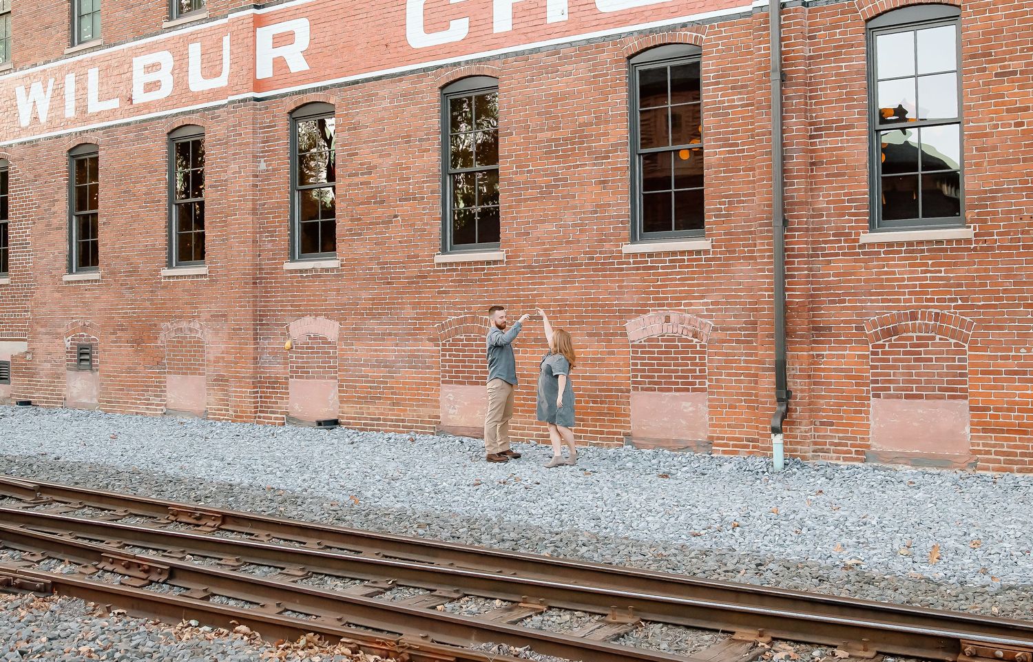 Two people stand beside a brick building with 'Wilbur Line' signage near railroad tracks on a gravel path.