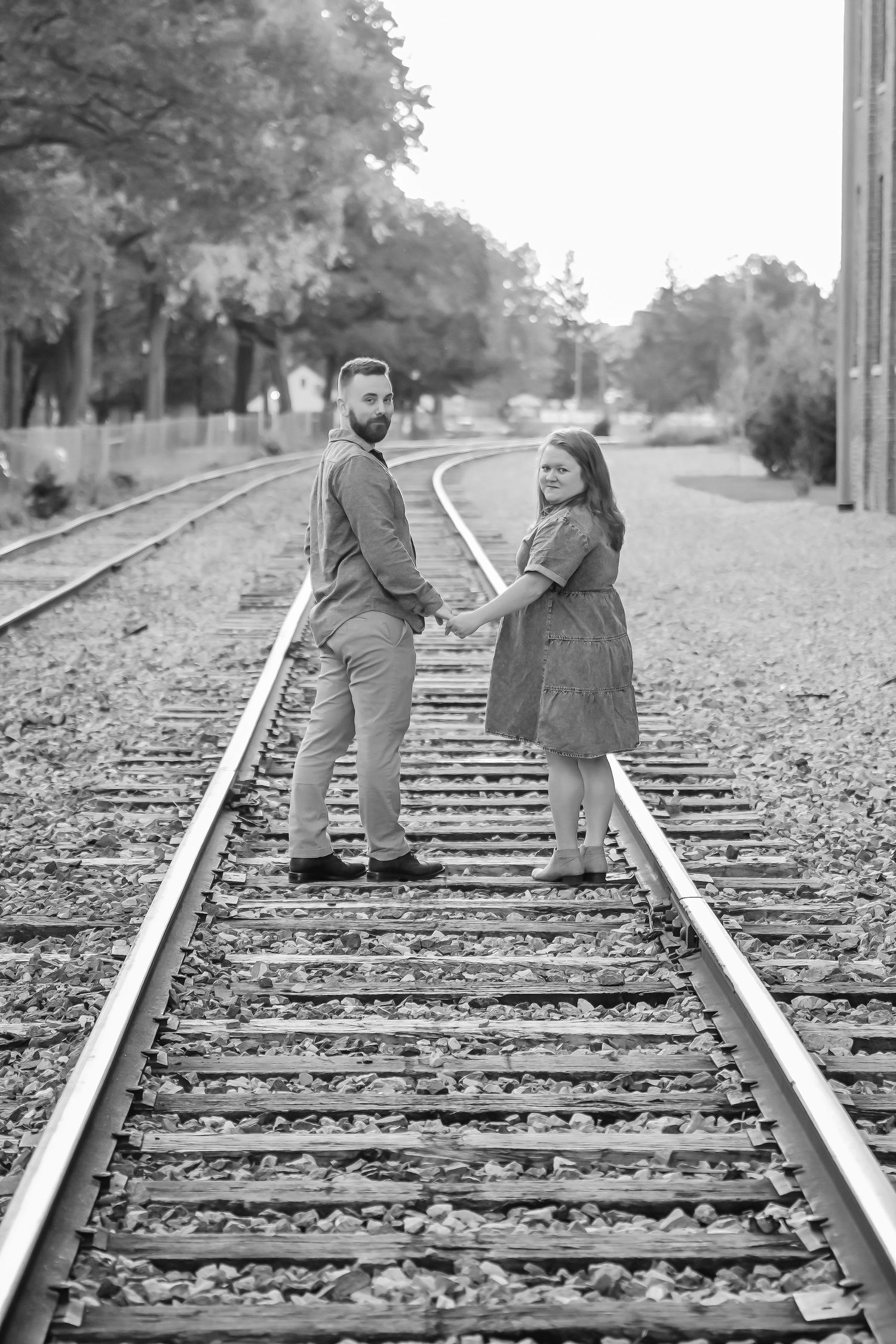 Black and white photo of a couple holding hands on railroad tracks, looking back at the camera.