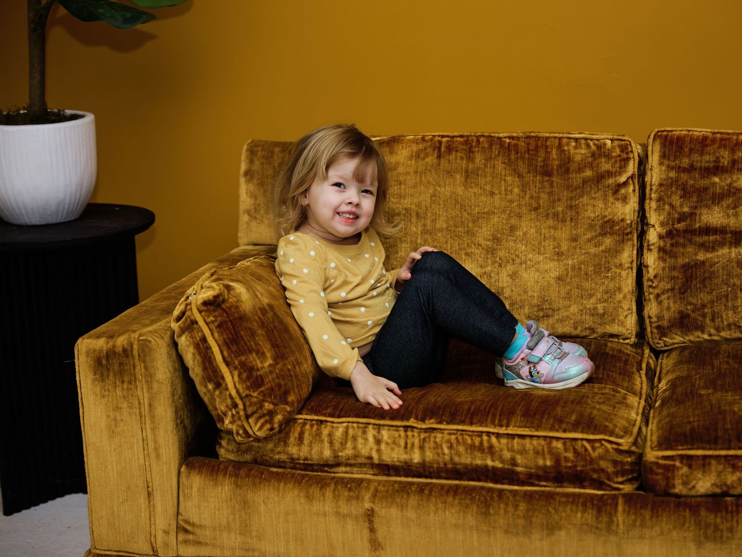 Young toddler girl sitting on a golden velvet sofa, wearing yellow top and dark jeans, smiling in a cozy room.