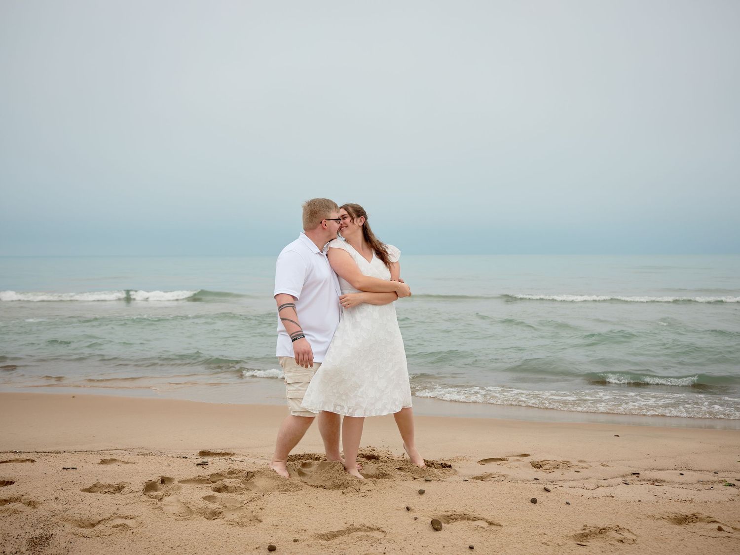 A romantic beachside moment with a couple embracing on the sandy shore as waves roll in on a misty day.