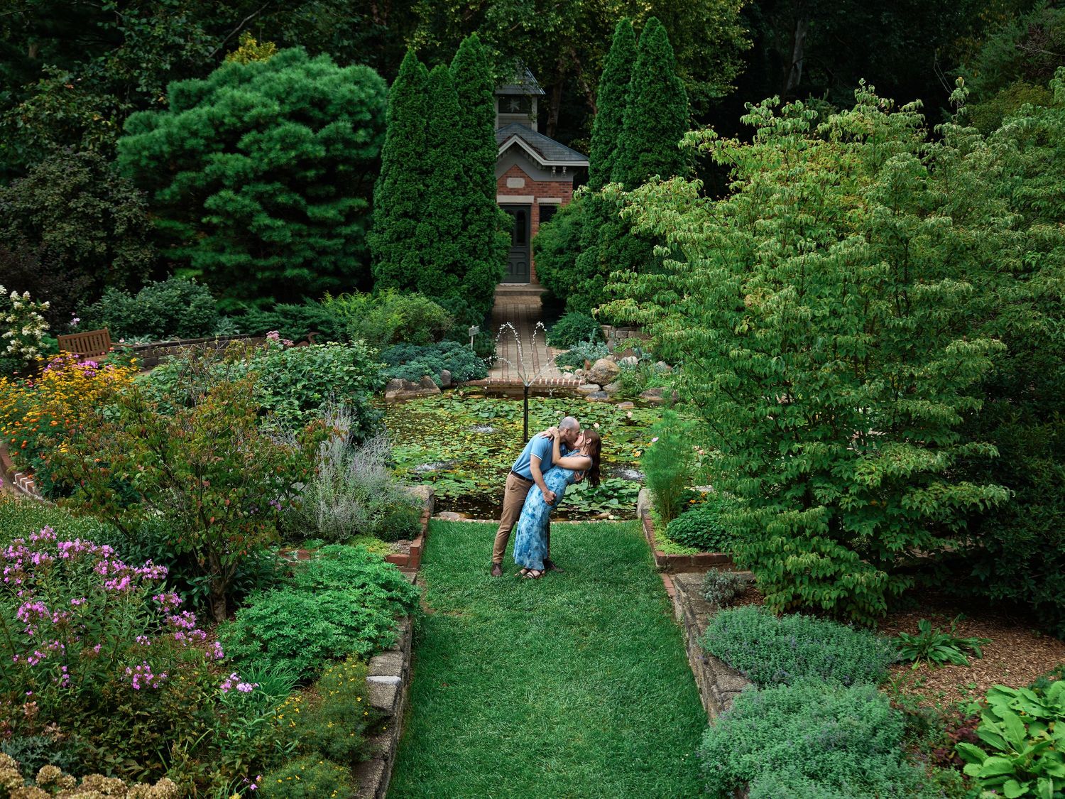 Couple embracing in a lush formal garden with manicured grass path, colorful flowers, and trees surrounding them.
