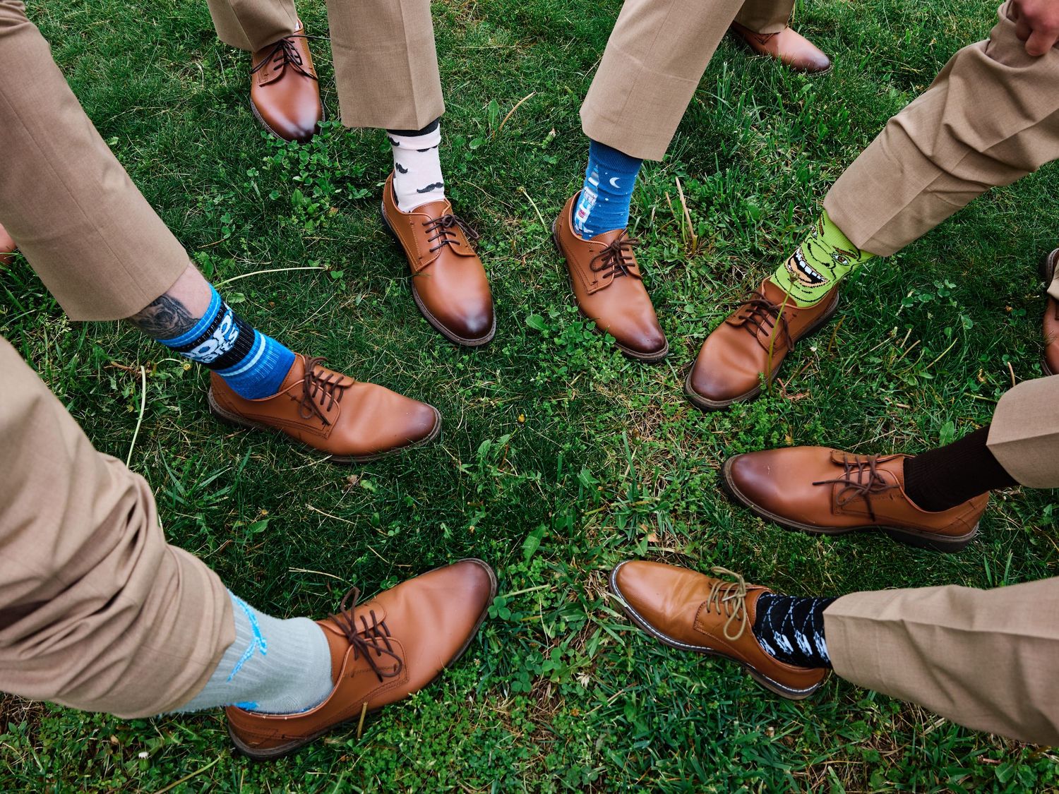 Groomsmen wearing colorful fun socks with brown oxford shoes and khaki pants standing in a circle on grass.