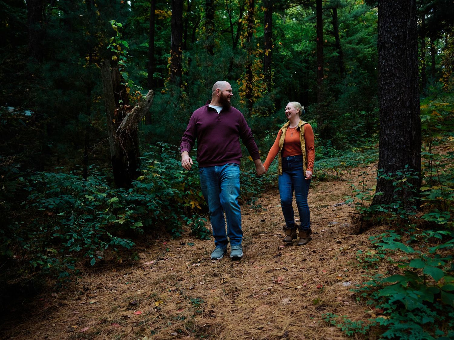 Two people hold hands while walking together on a dirt trail through a lush green forest.