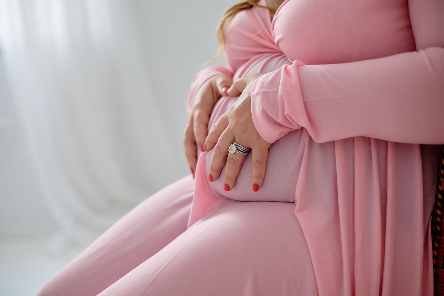 A pregnant person in a flowing pink dress cradles their baby bump against a white backdrop.