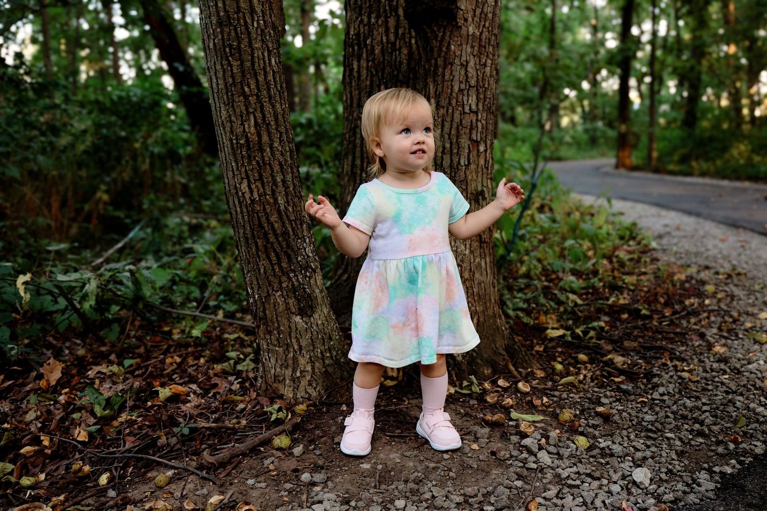 A small child wearing a pastel tie-dye dress stands between two trees on a wooded path.