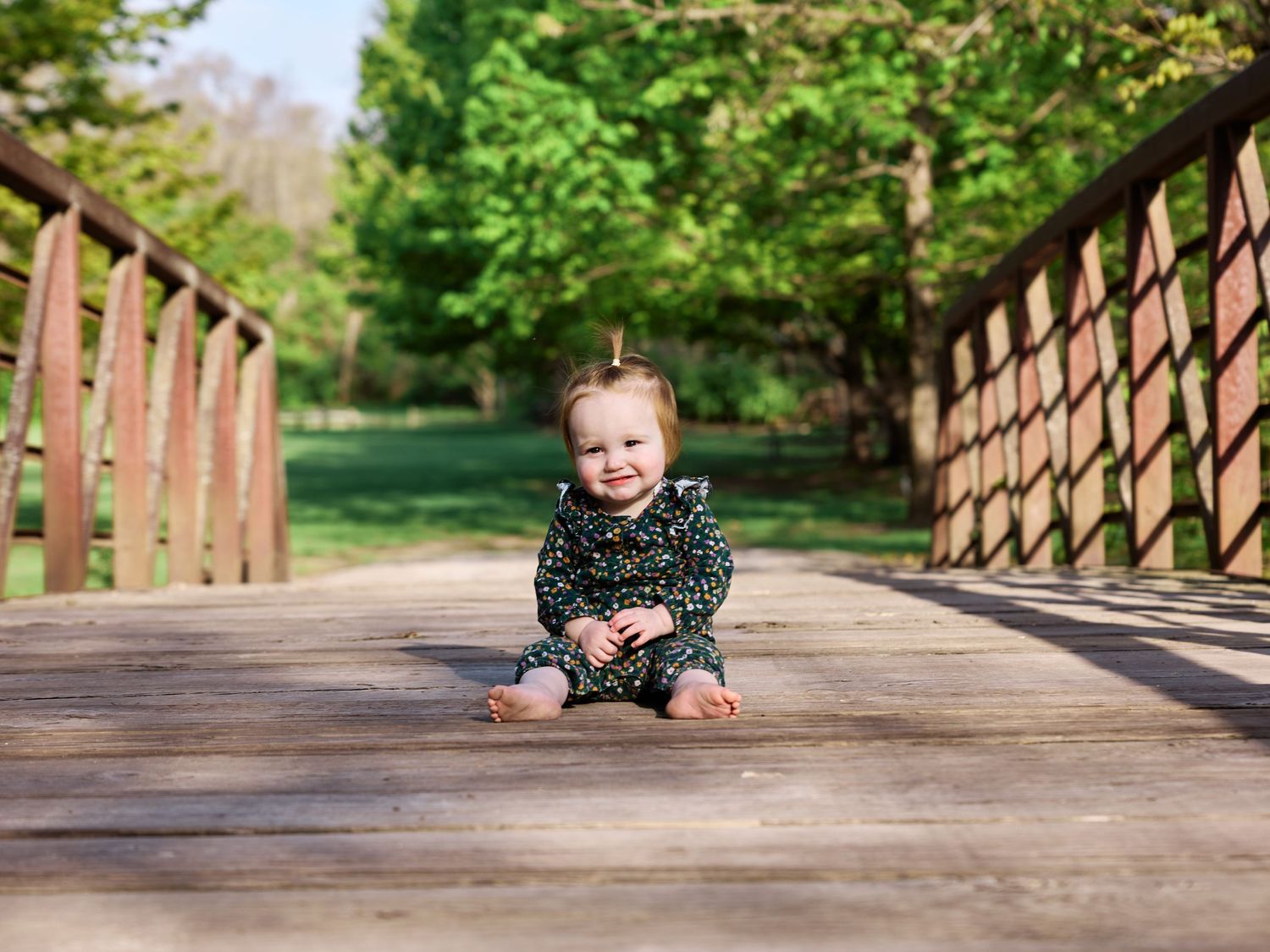Smiling baby sitting on a wooden bridge surrounded by lush green trees in a park.