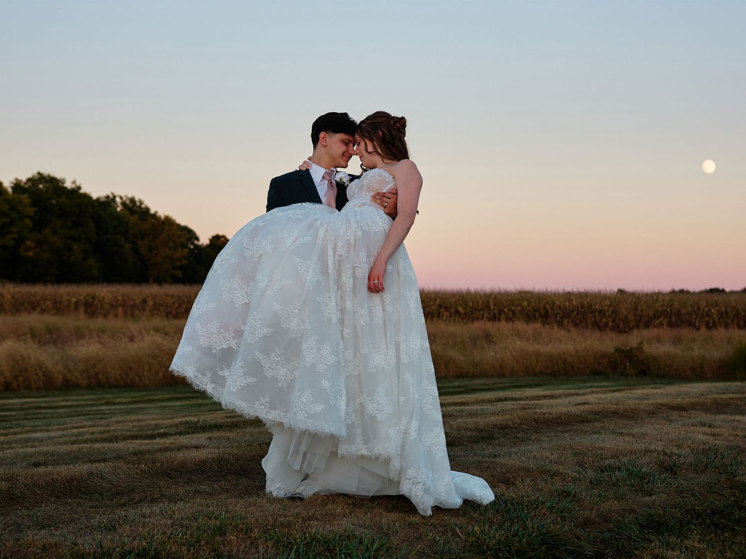 Groom lifting bride in lace wedding dress at sunset in a field with moon visible in pastel sky.
