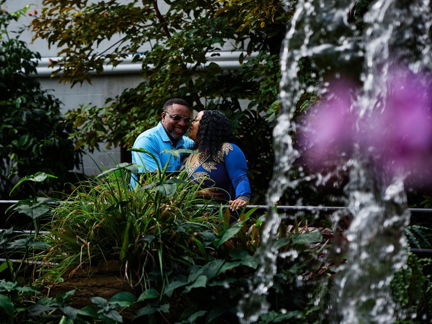 A couple shares a tender moment on a bench near a cascading fountain in a lush garden setting.
