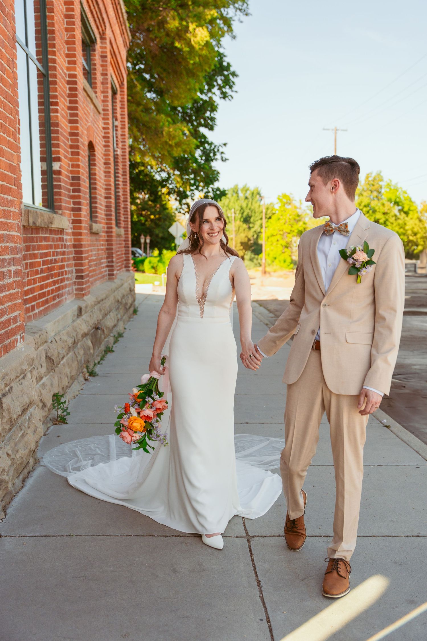 A bride in a white flowing dress and groom in a tan suit walk together along a brick building on a sunny day.