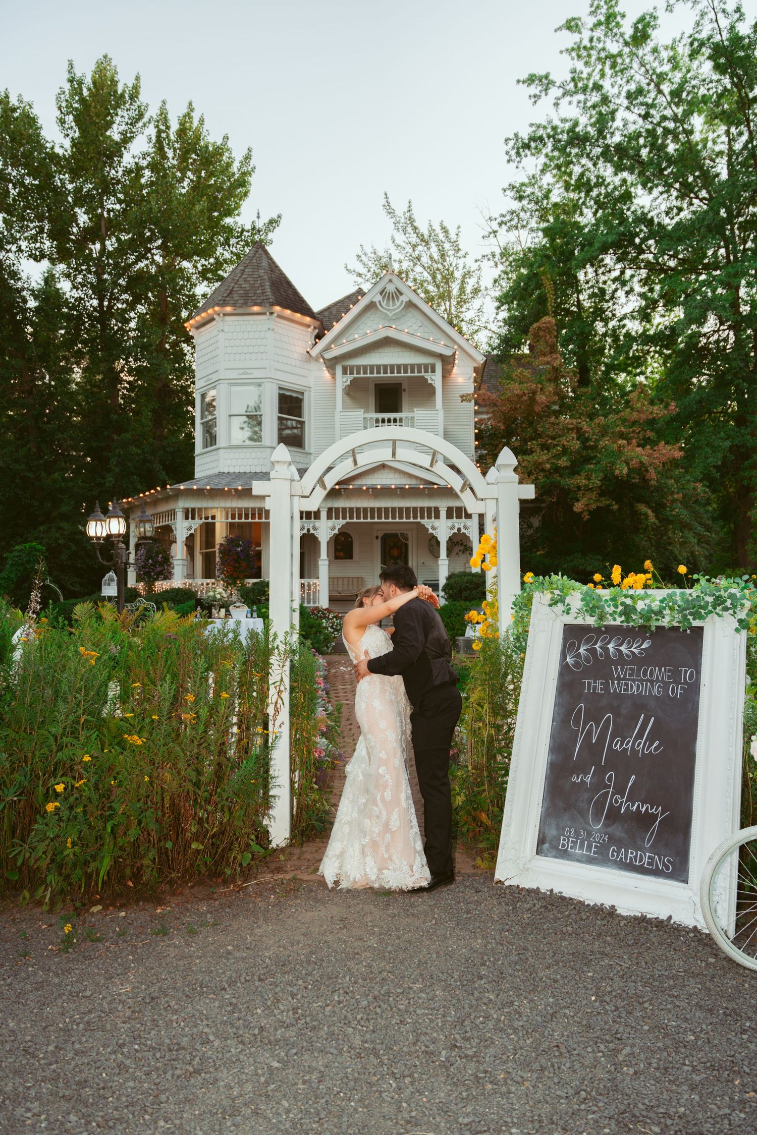 A romantic wedding moment outside a beautiful white Victorian mansion with a decorative chalkboard sign and garden.
