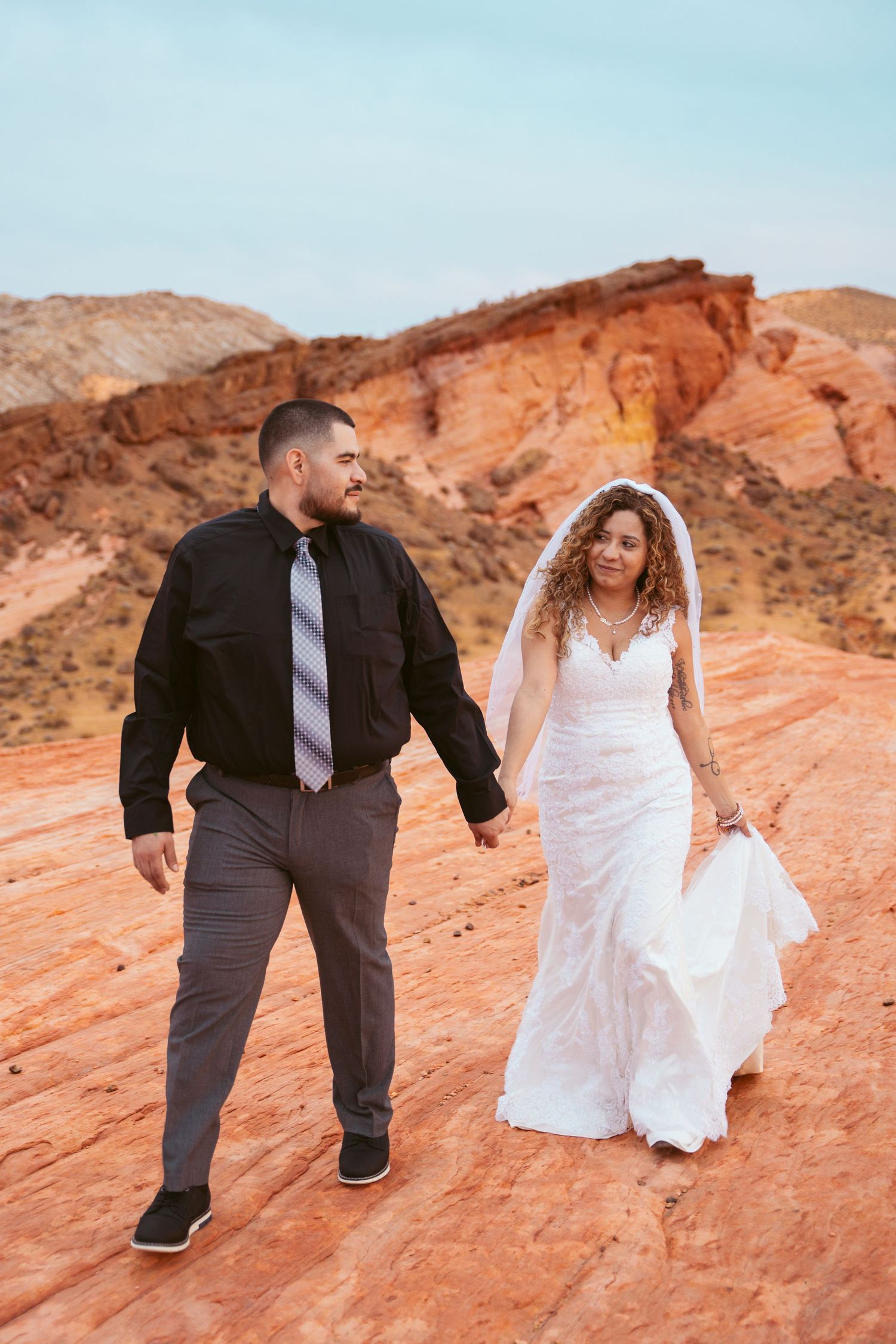 A couple in wedding attire playfully explores vibrant red rock formations in a desert landscape setting.