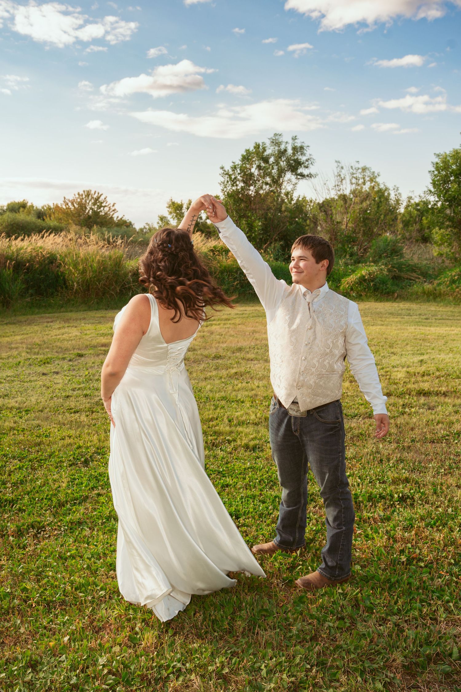 A romantic couple in wedding attire dances together in a sunny meadow during golden hour.