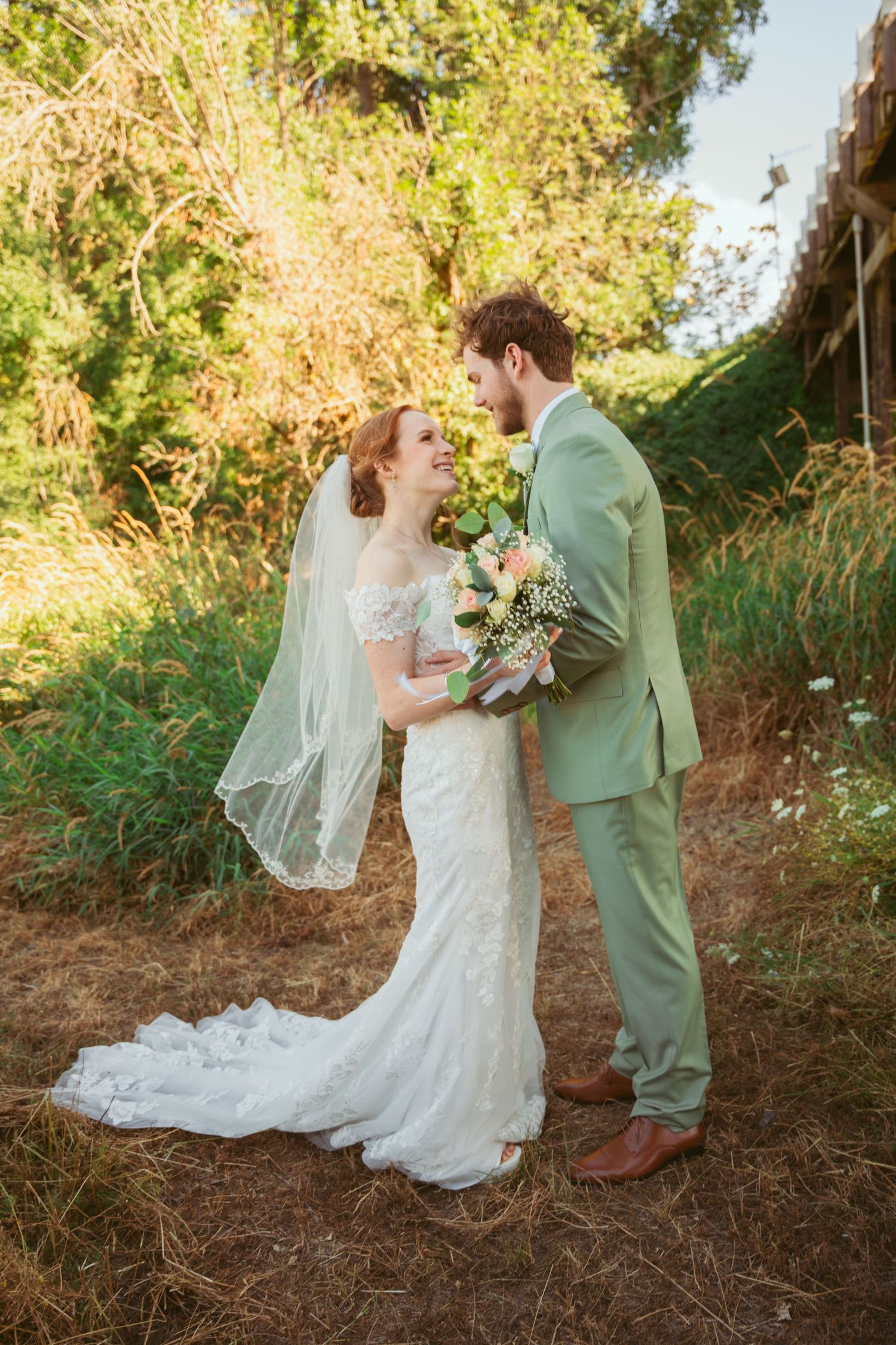 A couple in wedding attire embrace in a sun-dappled outdoor setting with lush greenery and a flowing white veil.