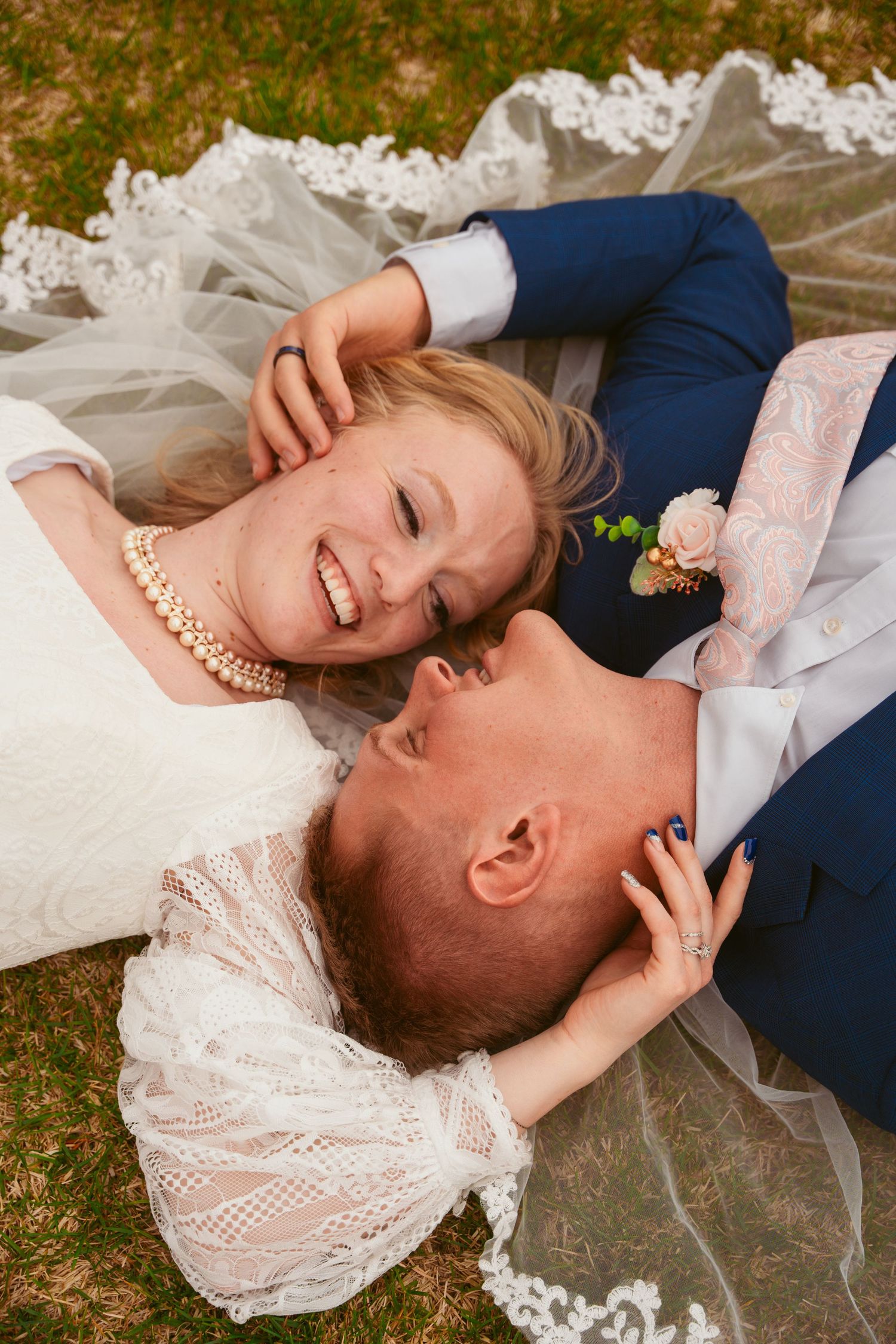 Happy couple laying together on grass wearing wedding attire with lace veil.