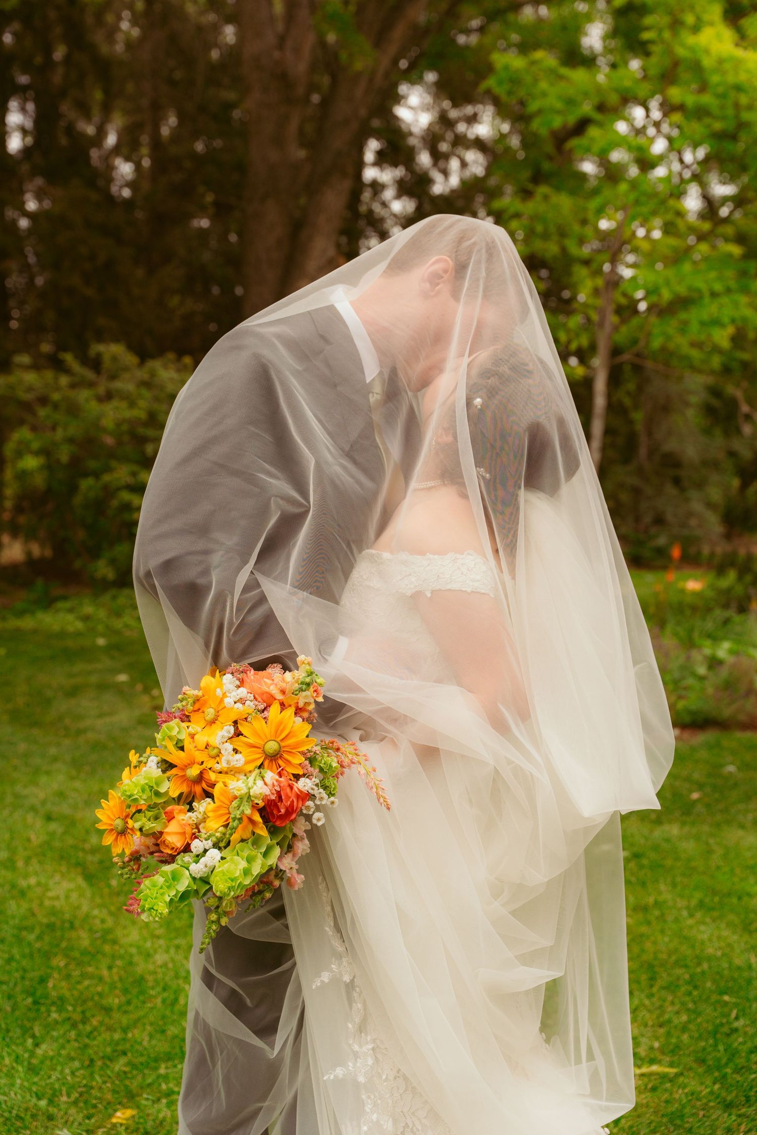 A bride in a white wedding dress covered by sheer veil holds vibrant orange and yellow autumn bouquet in a garden setting.
