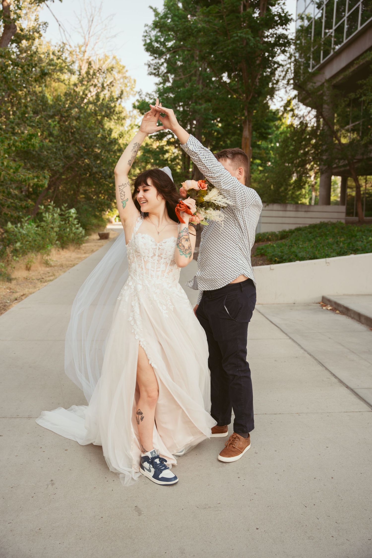A couple dances playfully in wedding attire, with the bride wearing a flowing white dress and groom in casual suit and sneakers.