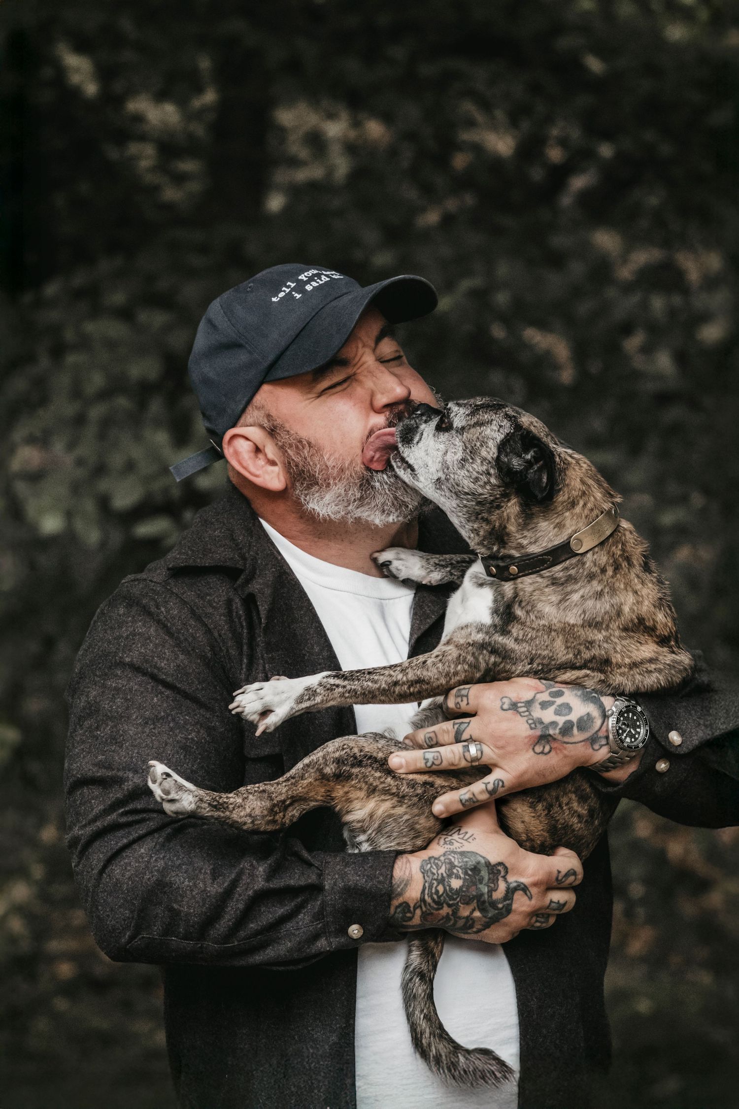 Tattooed man in cap being licked by brindle dog he holds in his arms outdoors.