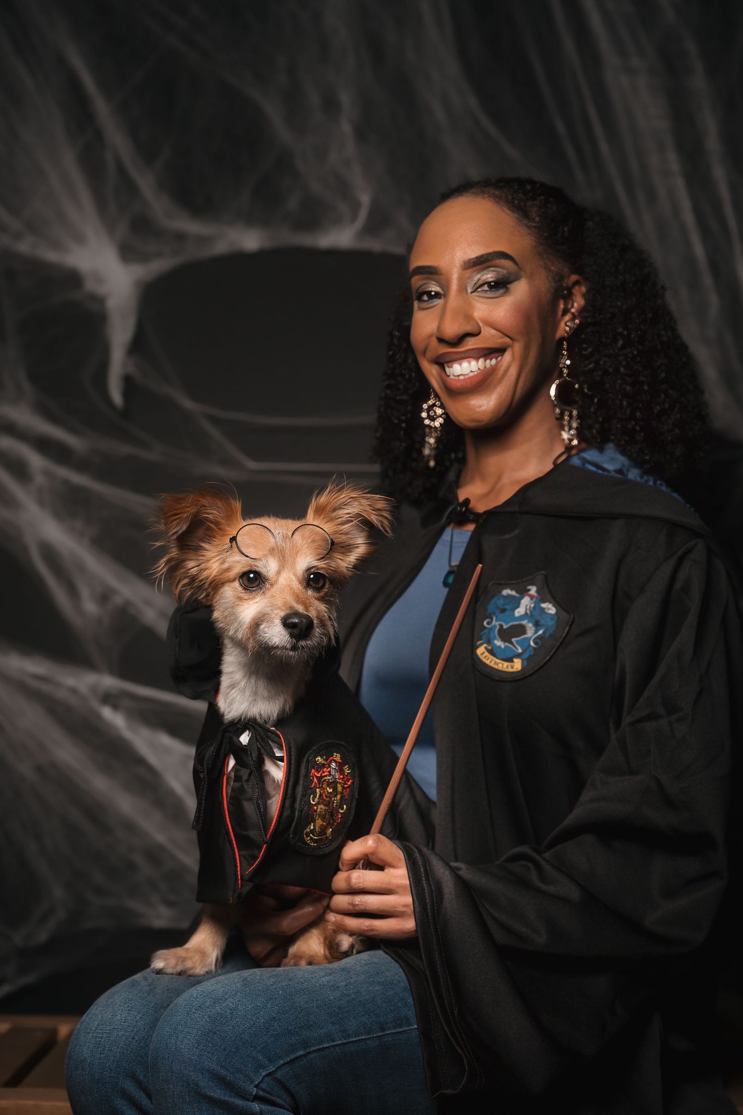 Smiling woman in a Ravenclaw-style wizard robe holds a small dog in round glasses and a matching robe against a spooky cobweb backdrop.