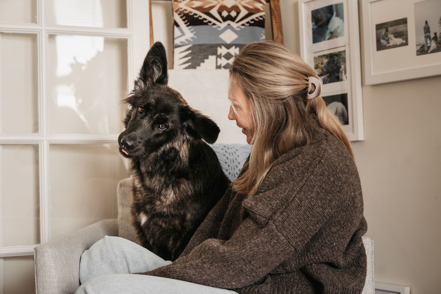 Blonde woman in brown sweater sitting with a dark German Shepherd dog indoors near a gallery wall.