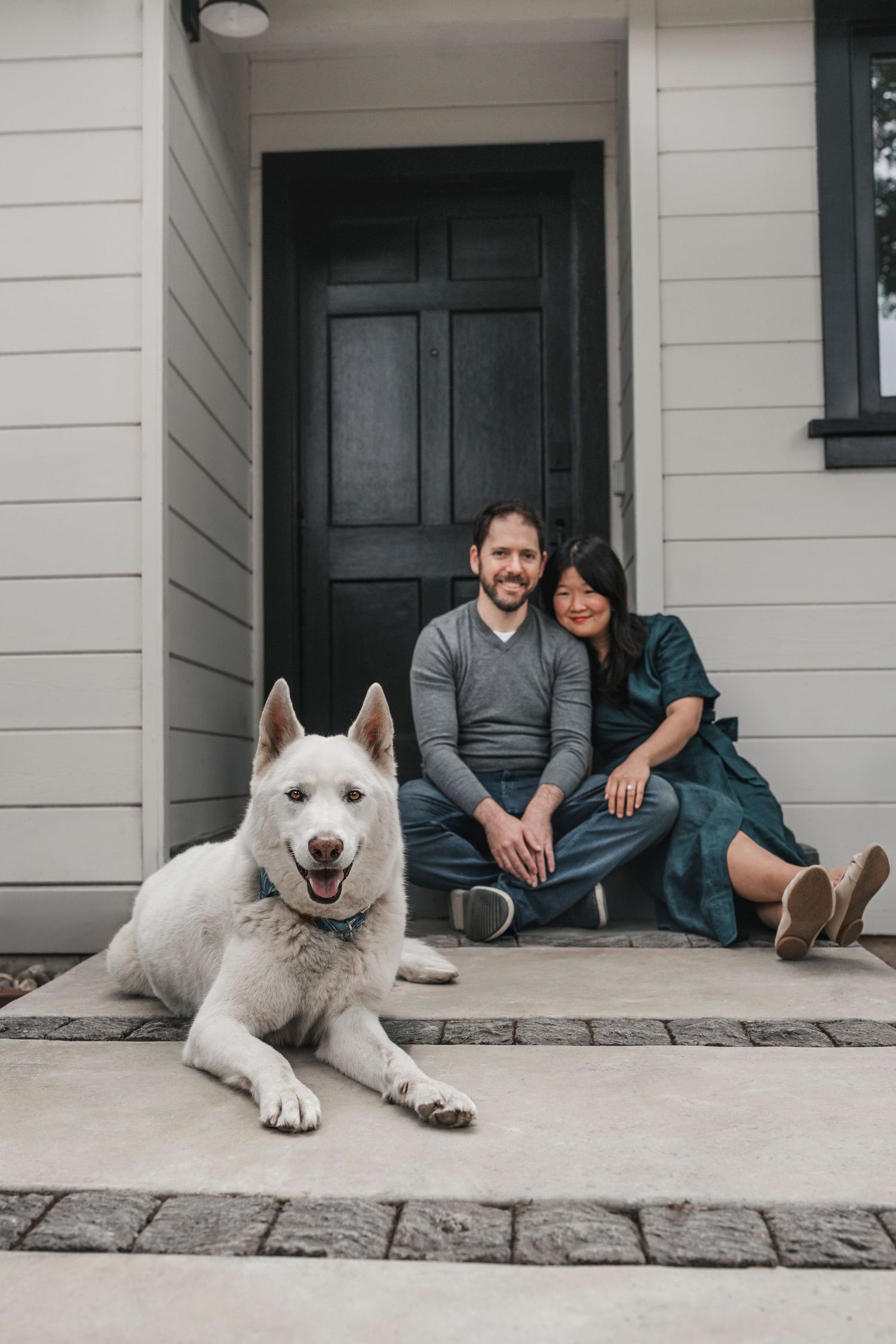 Couple sitting on front porch steps with white husky dog in foreground, smiling at camera by black door.
