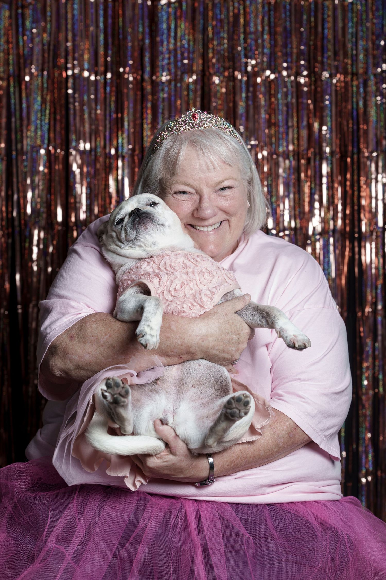 Smiling woman in pink tutu and tiara hugging her pug dressed in pink ruffles against sparkly backdrop.