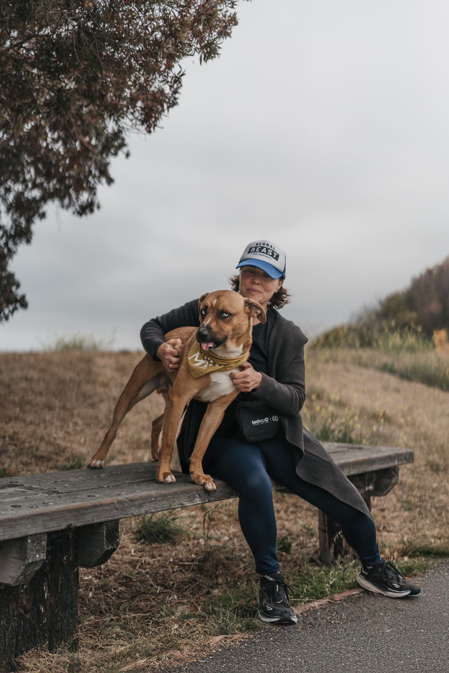 Woman in blue cap hugging brown Pit Bull mix dog on wooden bench in scenic outdoor setting.
