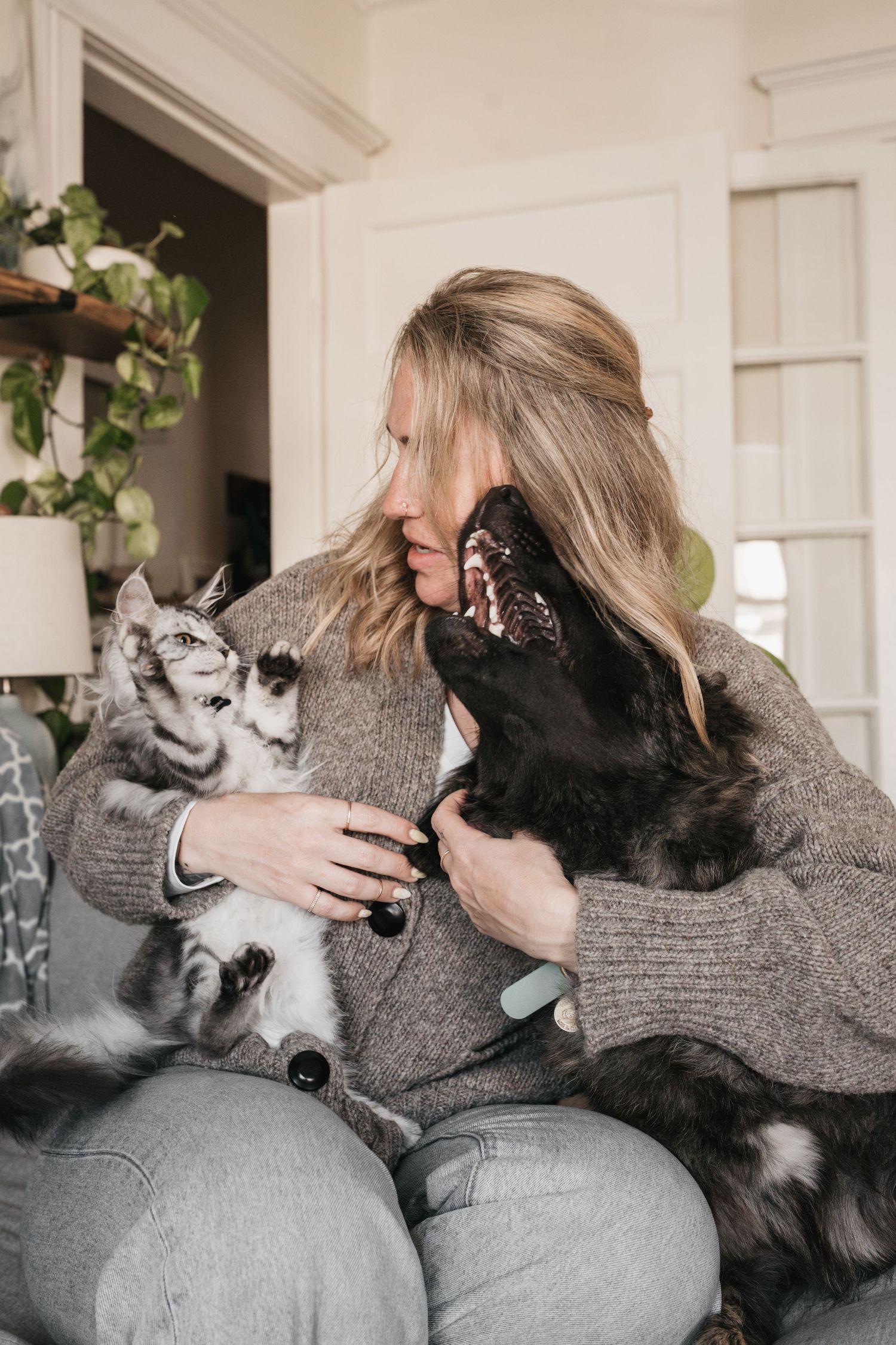 Blonde woman cuddling a black dog and maine coon cat on a cozy couch indoors.
