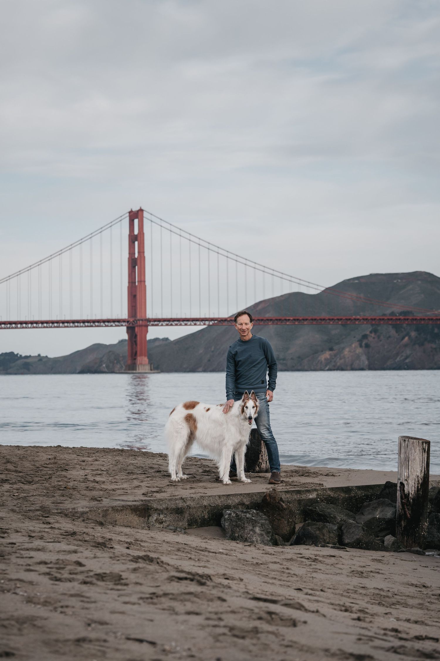 Man and dog standing on beach with Golden Gate Bridge in background in San Francisco, CA.
