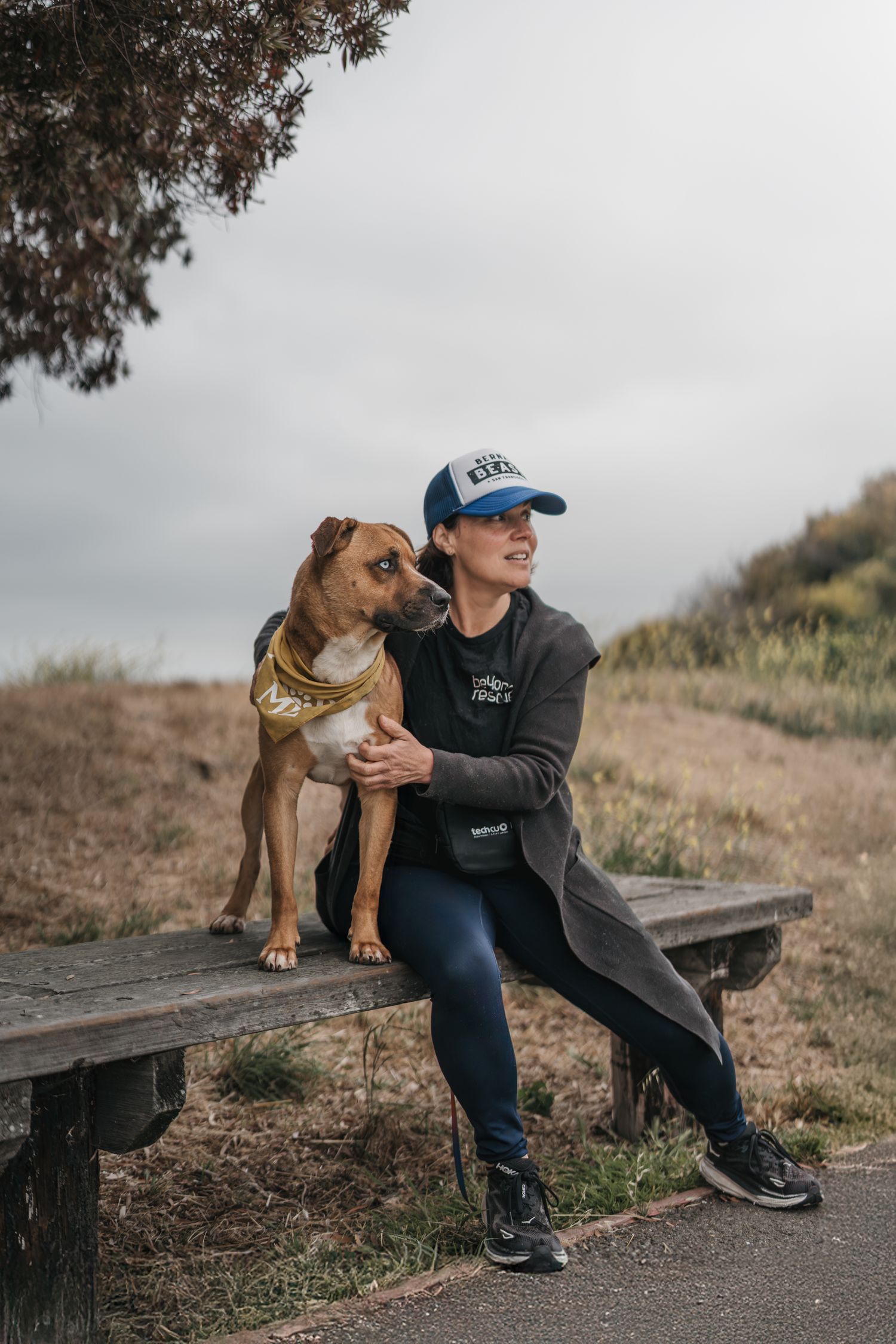 Woman in blue cap sitting on wooden bench with brown Pit Bull mix dog wearing gold bandana outdoors.