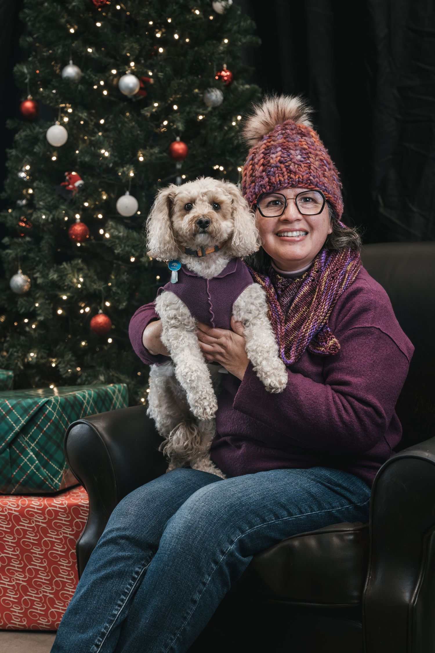 Smiling woman in purple sweater and knit hat holds a small white dog in front of a decorated Christmas tree.