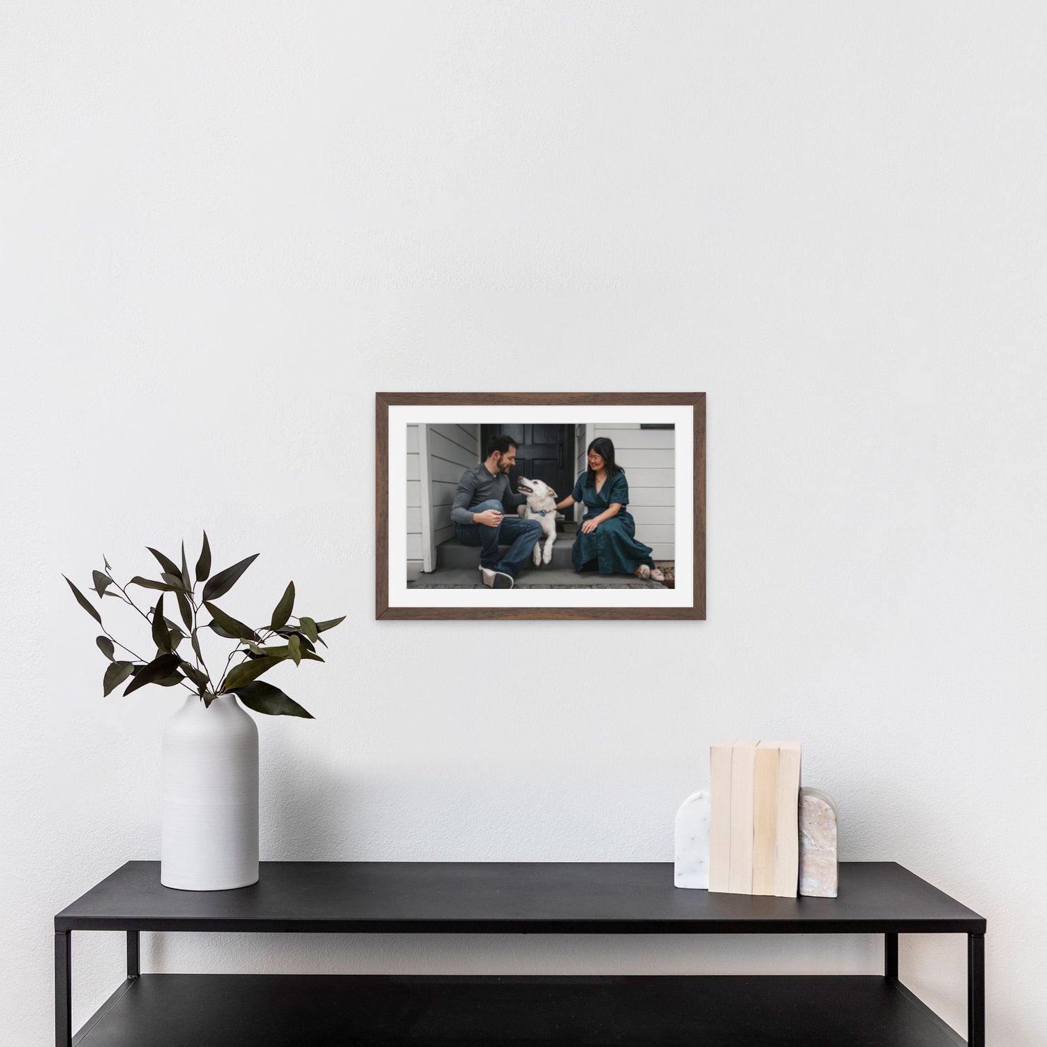Couple with dog in walnut frame hanging above black console table with plant and books.