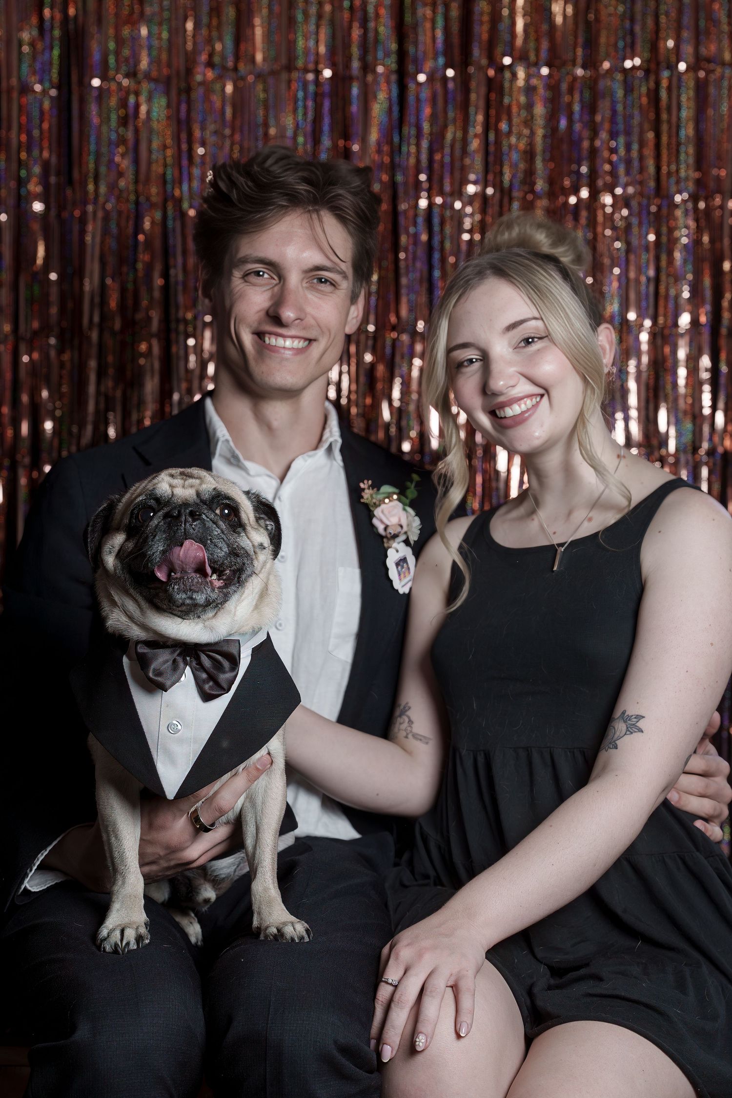 Couple in formal attire smiling with a pug dog wearing a tuxedo in front of a shiny backdrop. At the first annual pug prom.