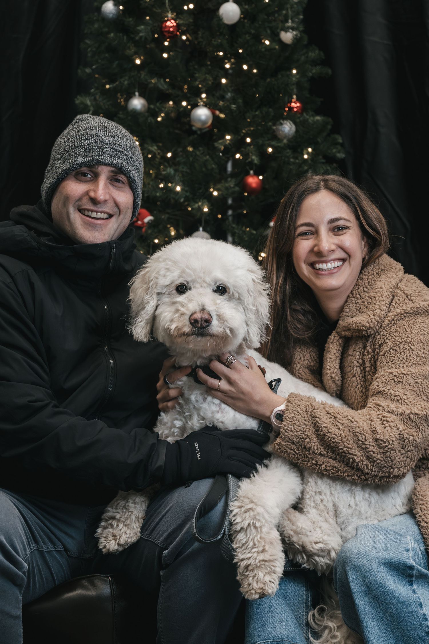 Smiling couple holding a white fluffy dog in front of a decorated Christmas tree with warm lights.