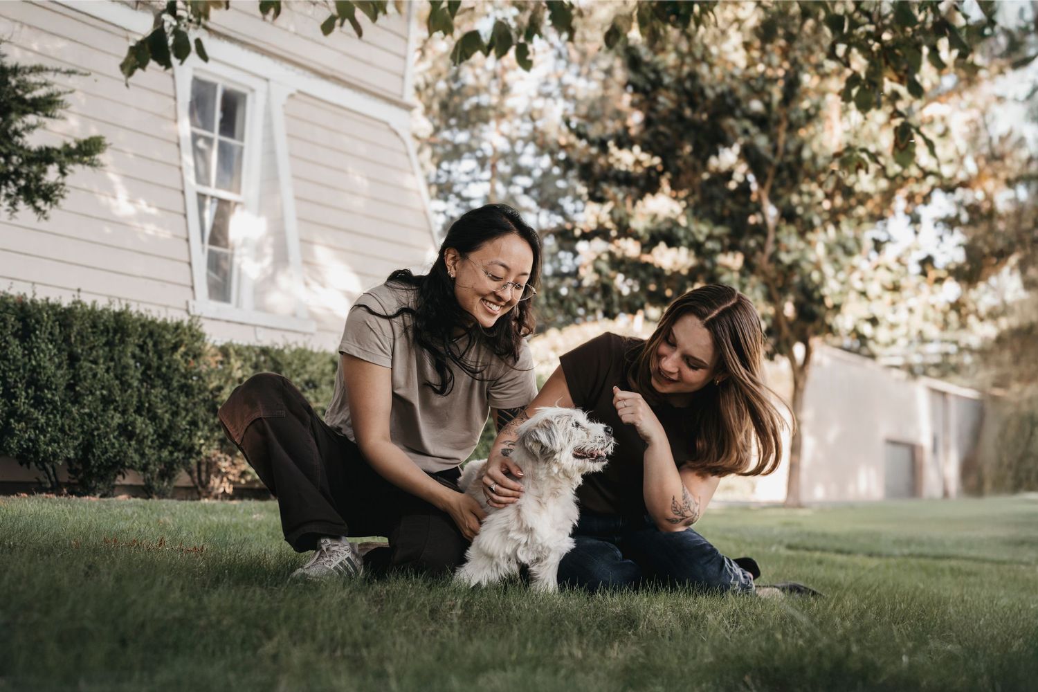 Two women sitting on grass in a backyard, petting and playing with a small white fluffy dog.