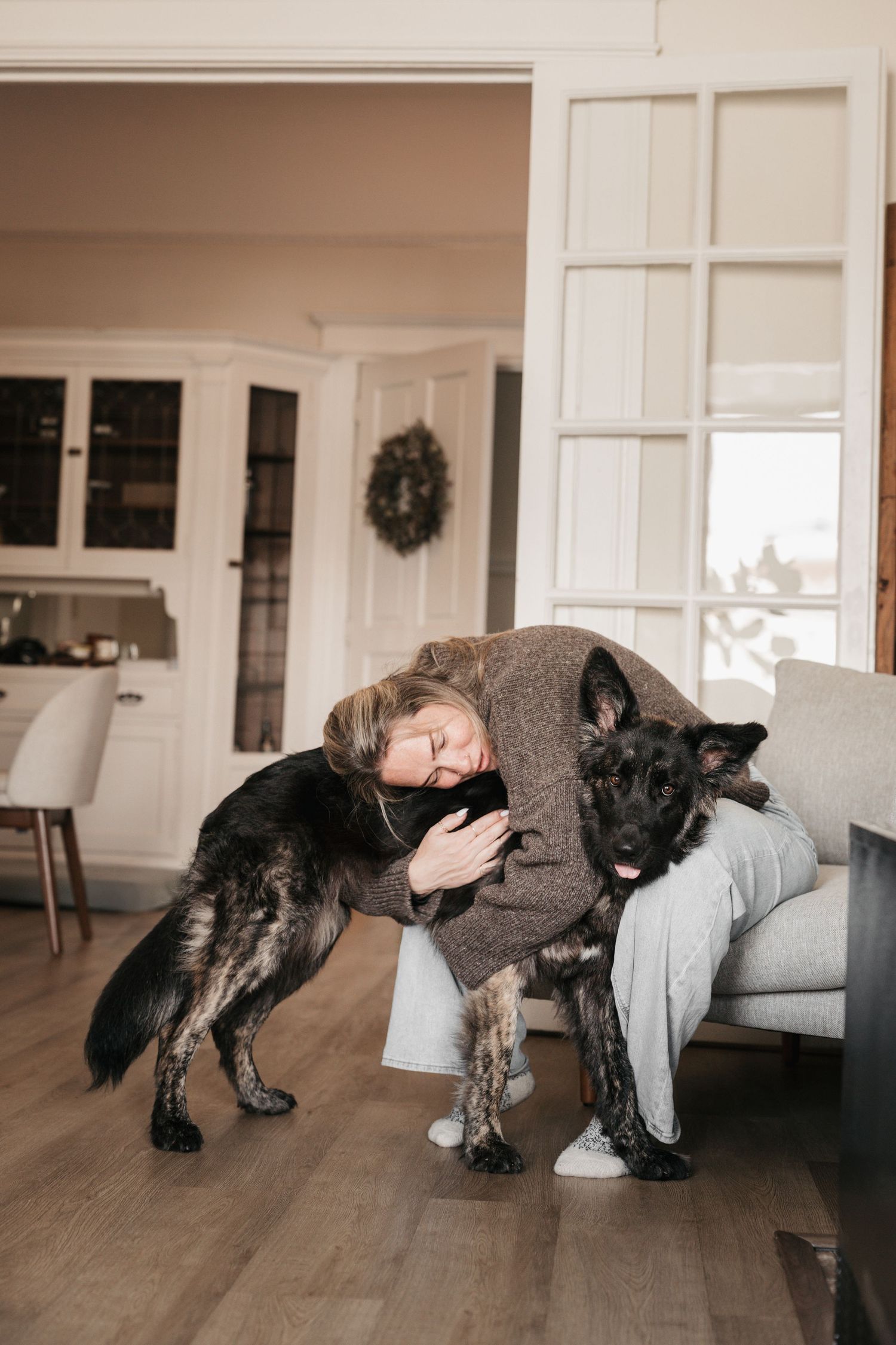 Woman in brown sweater hugging a large dark German Shepherd dog in a bright living room.