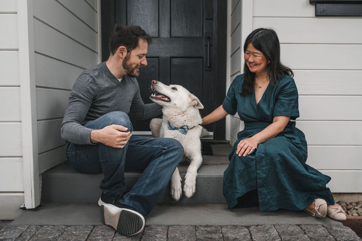 A couple sits on porch steps laughing with their happy white Labrador dog between them.
