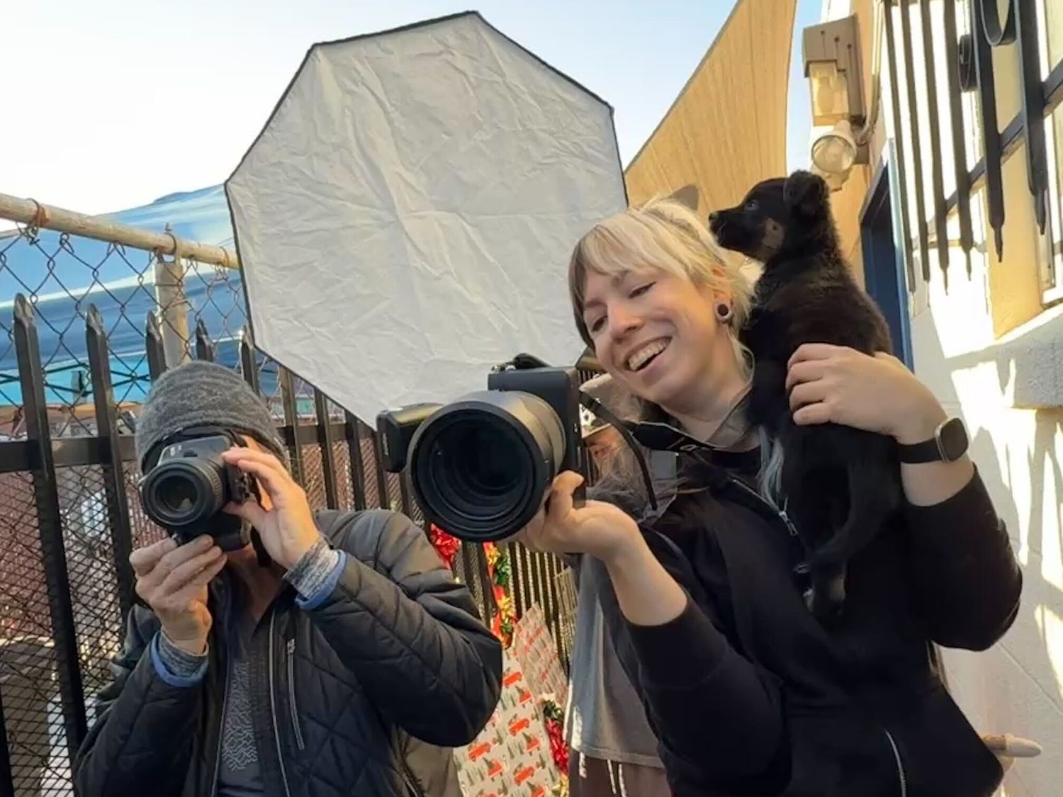 Two photographers with cameras and a small puppy on one's shoulder shoot outdoors near a softbox light.