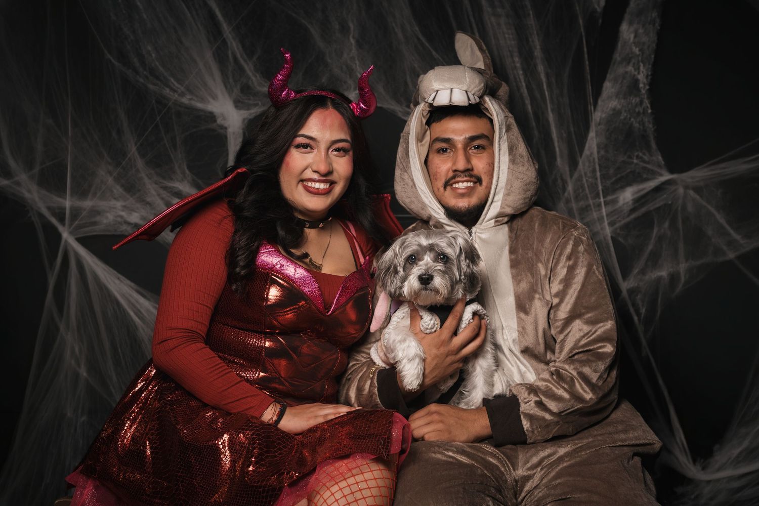 Smiling couple in Halloween costumes, a devil and a hooded creature, holding a small gray dog against a spooky spiderweb backdrop.
