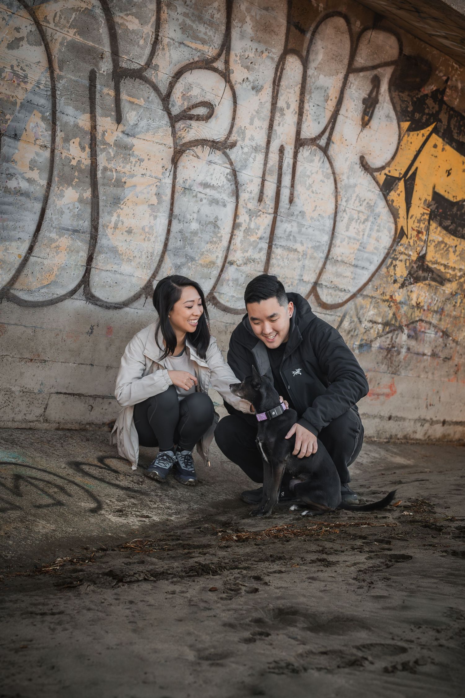 A couple crouches with their black dog in front of a graffiti wall on a sandy urban surface at Fort Funston San Francisco