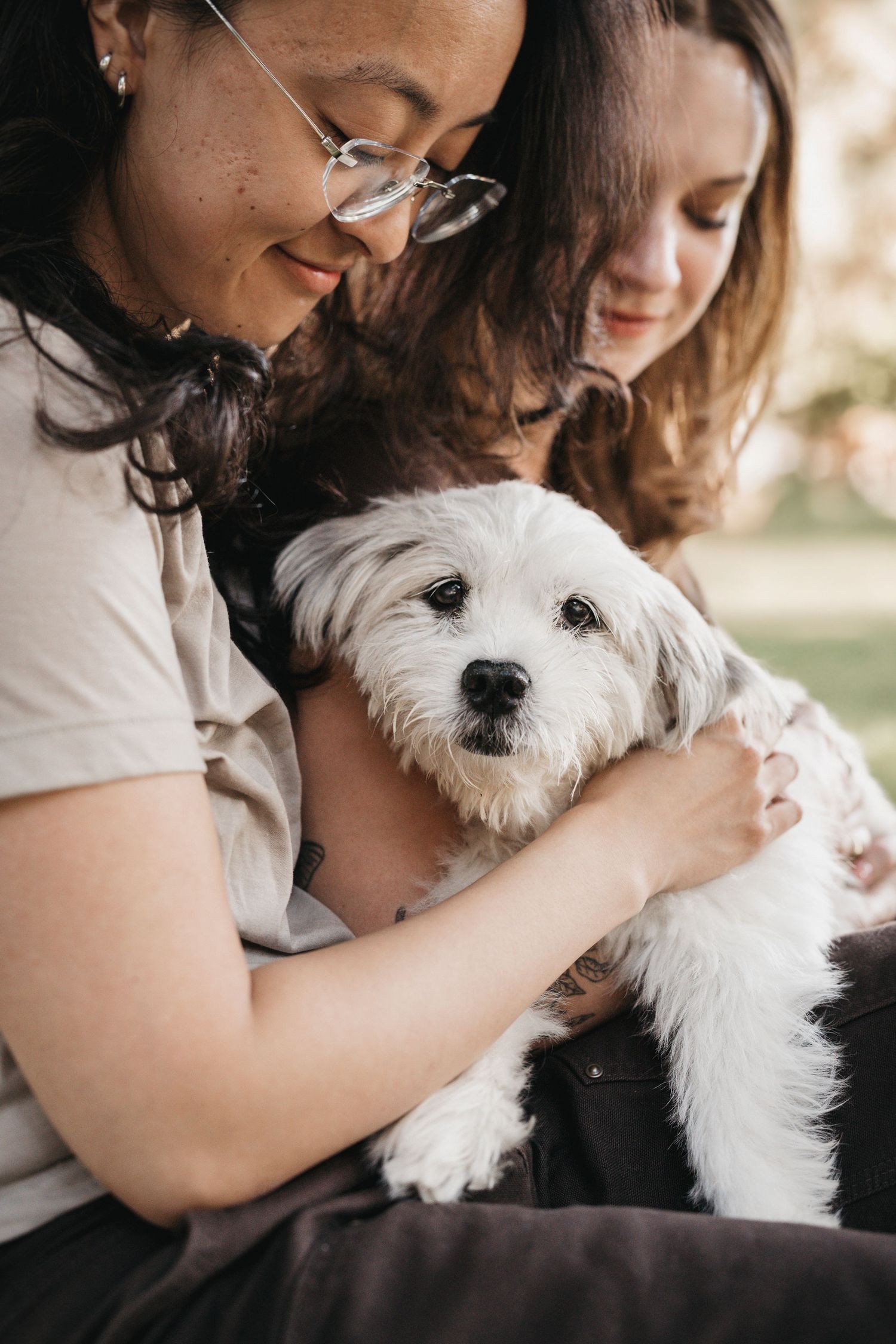 Two young women smiling and cuddling a fluffy white dog outdoors in a park setting.