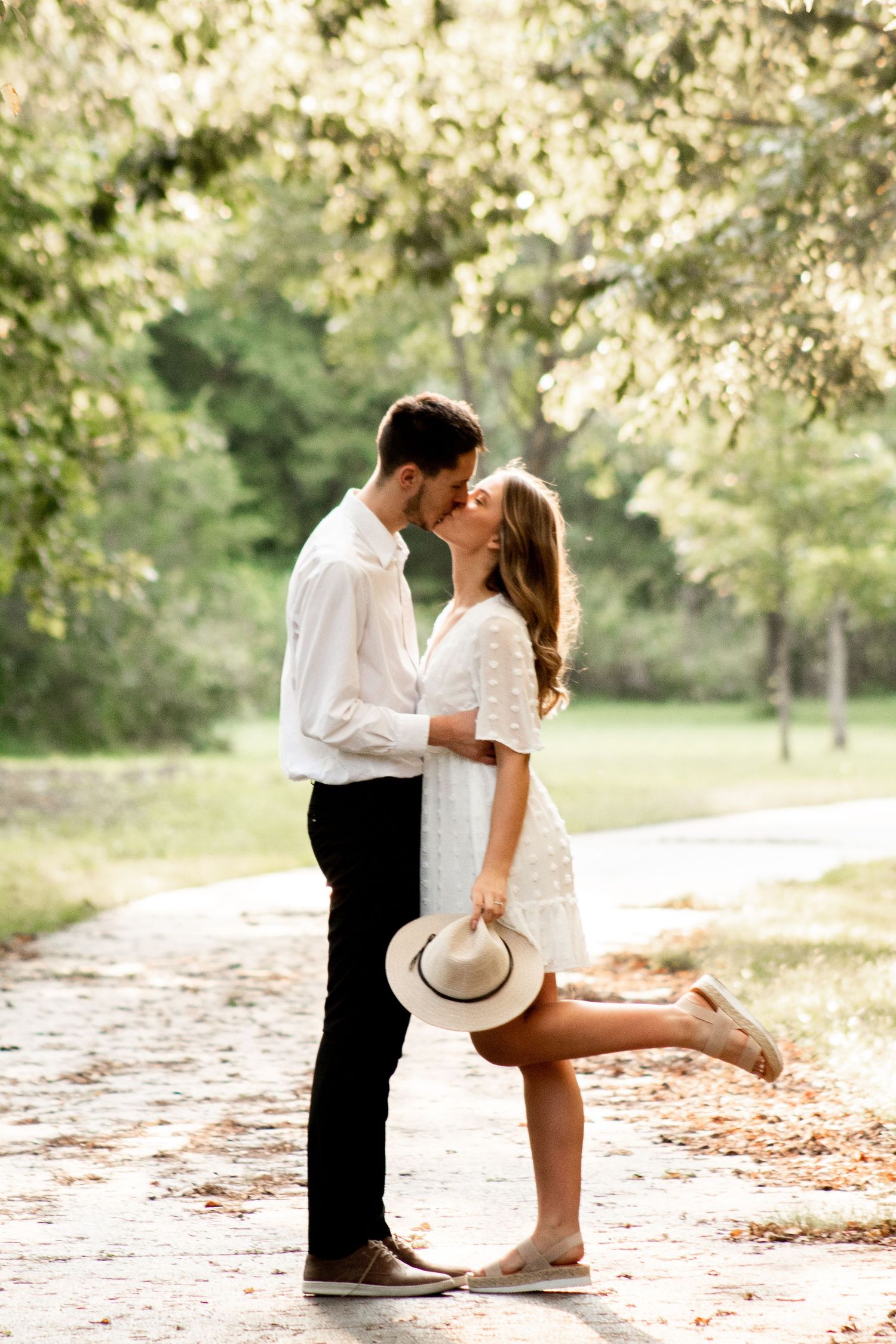 Rom-com style kiss with a sunlit background, the fiancés sharing a kiss, with her foot popped and one hand holding her hat as it falls.
