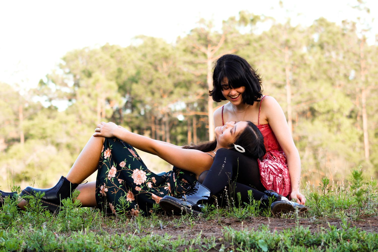 Friends lying in a grassy field: one sitting criss-cross, looking down, while the other lays her head in her lap, smiling up, with her body