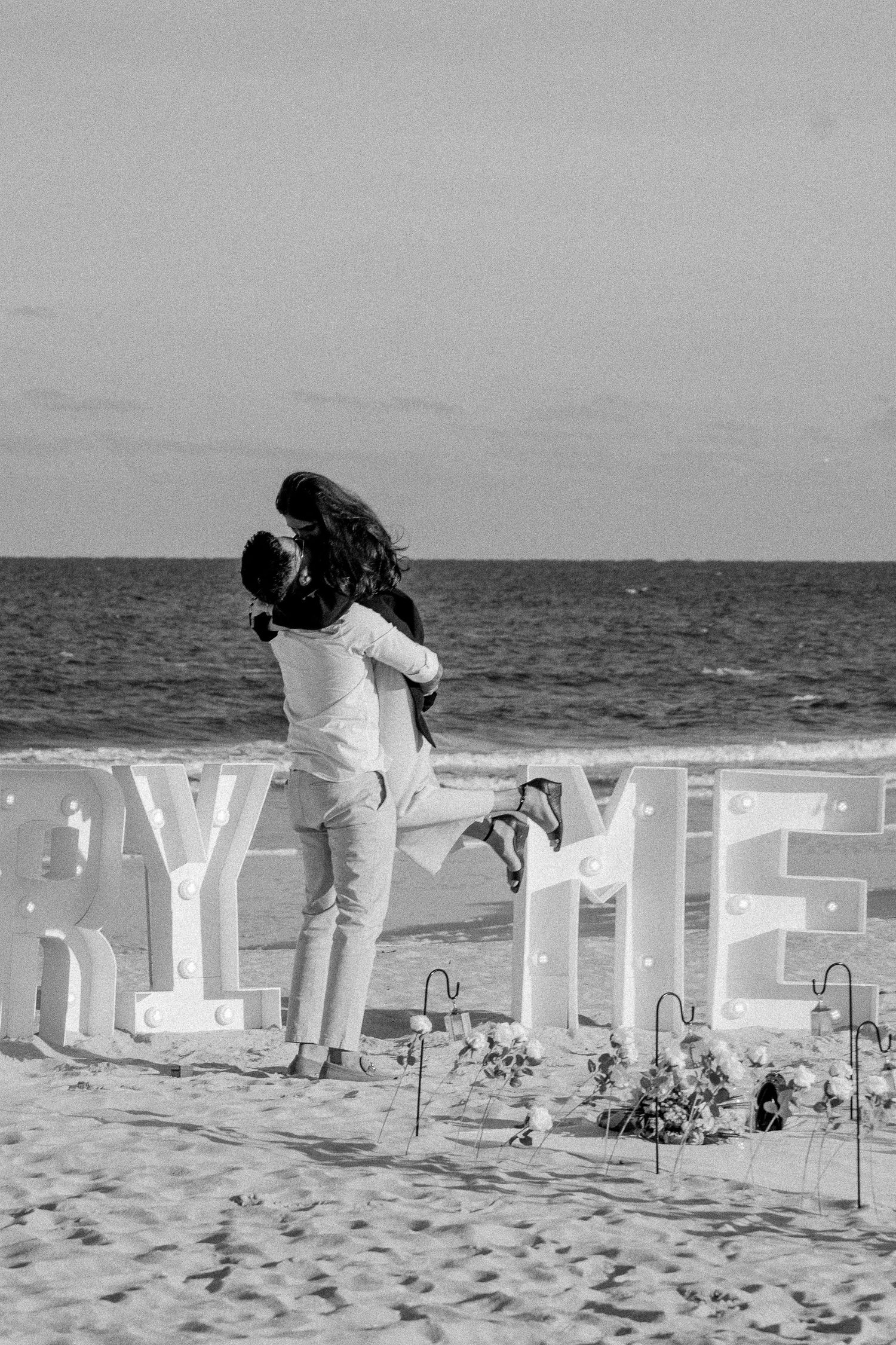 Black-and-white photo of a man joyfully lifting his fiancée into the air after she said yes to his marriage proposal, capturing their excite