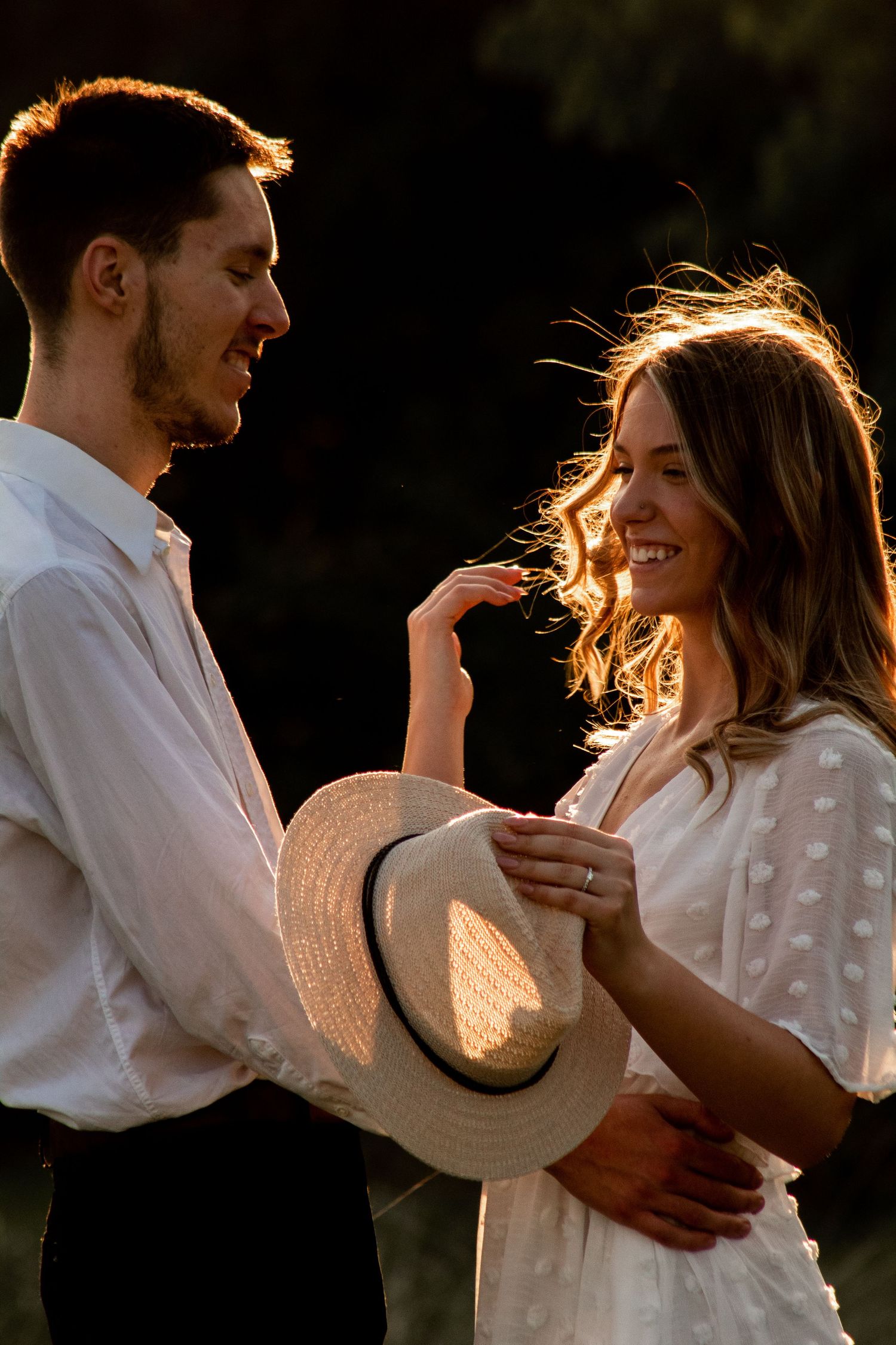 Engagement shoot at sunset, girl holding her hat as her fiancé wraps his hands around her waist, sun highlighting her hair from behind.