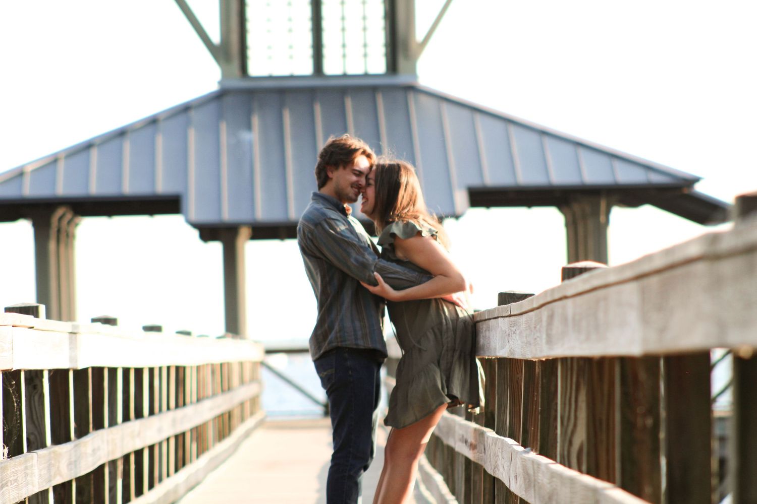 Engagement photo on a bridge, woman leaning into fiancé for support, holding his arms as he buries his face in hers and pushes her back.
