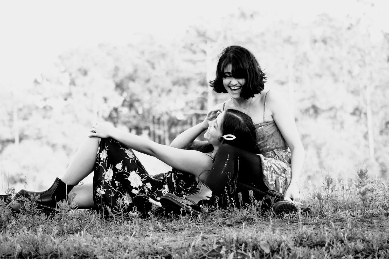 Friends lying in a grassy field: one sitting criss-cross, looking down, while the other lays her head in her lap, smiling up, with her body