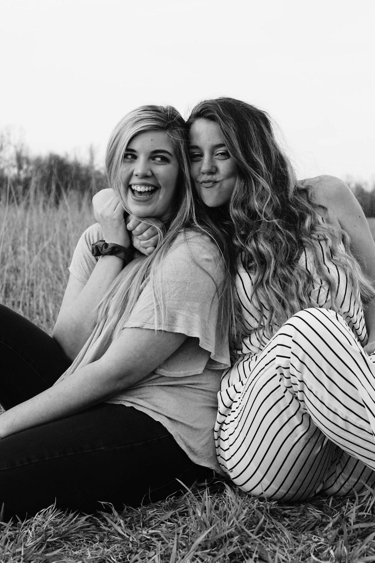 Two sisters sitting together in a field, one with her arm wrapped around the other's neck, pulling her in close as