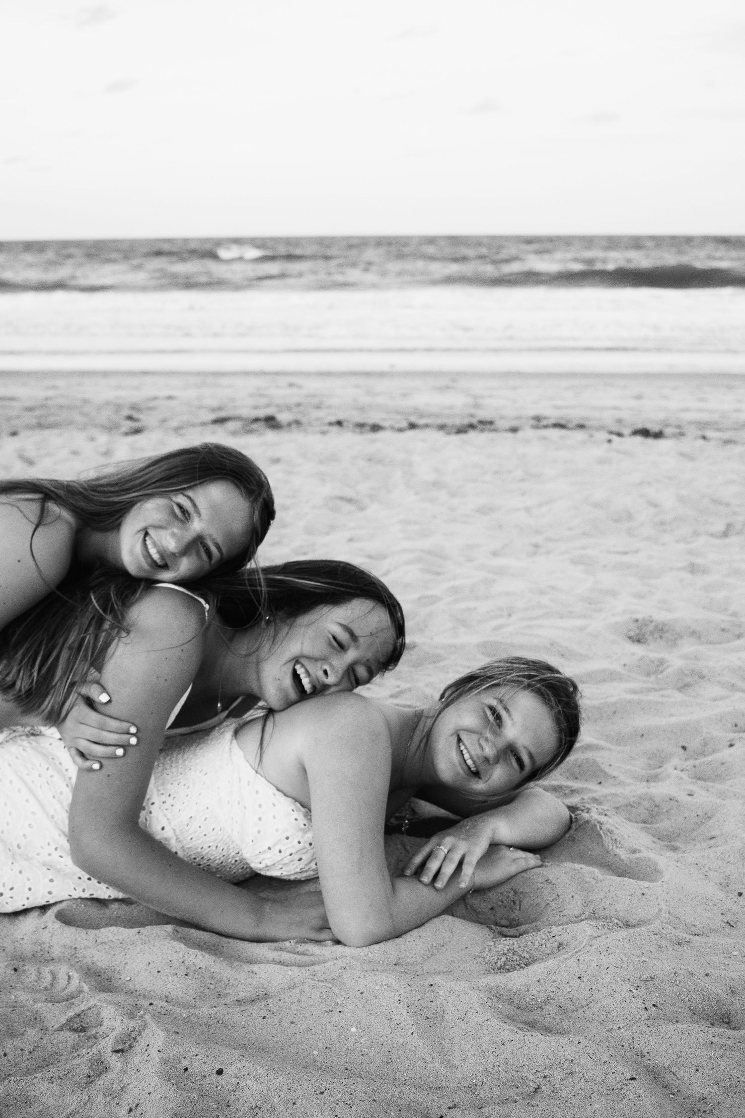 Beach photo of three girls lying on top of each other, laughing and smiling together.